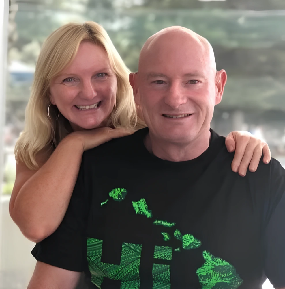 Smiling couple posing together indoors, with woman resting her arms on man’s shoulders while both face the camera in casual black clothing