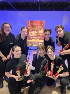 Group of theatre performers celebrating with trophies at the Shire Theatre Competition after receiving awards on stage.